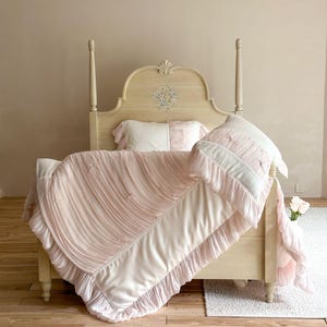 May include: A cream-colored wooden bed frame with a floral motif on the headboard, featuring a light pink and white bedding set. The comforter and pillows are adorned with vertical ruffles, small fabric bows, and a smooth, solid-colored border.