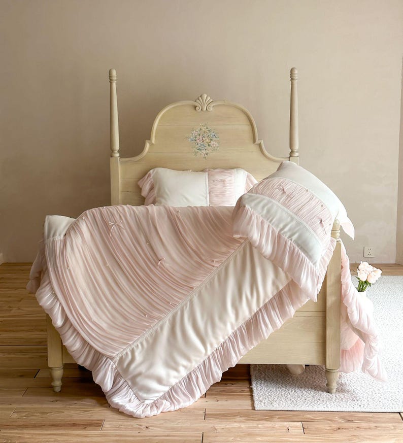 May include: A cream-colored wooden bed frame with a floral motif on the headboard, featuring a light pink and white bedding set. The comforter and pillows are adorned with vertical ruffles, small fabric bows, and a smooth, solid-colored border.