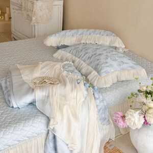 May include: A light blue bedding set featuring a quilted coverlet with a ruffled cream-colored skirt, two matching ruffled pillows, and a sheer cream-colored fabric overlay draped across the bed, accented by a circular pearl-like decorative piece.