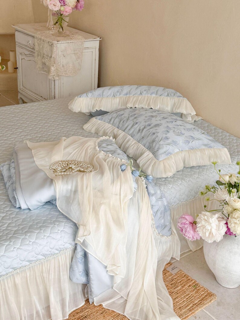 May include: A light blue bedding set featuring a quilted coverlet with a ruffled cream-colored skirt, two matching ruffled pillows, and a sheer cream-colored fabric overlay draped across the bed, accented by a circular pearl-like decorative piece.