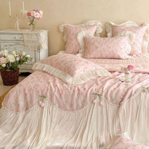 Puede incluir: Un juego de cama de tela rosa claro con un patr&oacute;n repetitivo de tulipanes. El edred&oacute;n y las fundas de almohada est&aacute;n adornados con bordes de volantes blancos y detalles decorativos de tulipanes de tela a lo largo de los bordes.