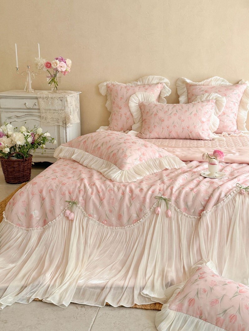 Puede incluir: Un juego de cama de tela rosa claro con un patr&oacute;n repetitivo de tulipanes. El edred&oacute;n y las fundas de almohada est&aacute;n adornados con bordes de volantes blancos y detalles decorativos de tulipanes de tela a lo largo de los bordes.