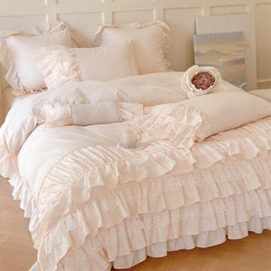 May include: A blush-colored bedding set with ruffled trim and lace accents. The set includes a duvet cover, pillowcases, and a decorative bolster pillow. A small teacup and saucer sit on the bed, adding a touch of elegance to the scene.