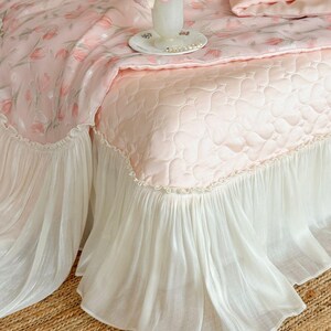 Puede incluir: Una colcha acolchada de color rosa claro con un patr&oacute;n de corazones repetido y un fald&oacute;n blanco con volantes. La ropa de cama est&aacute; adornada con una manta con estampado floral en tonos rosa y verde, junto a un jarr&oacute;n blanco con una flor rosa.