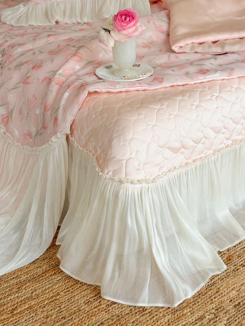 Puede incluir: Una colcha acolchada de color rosa claro con un patr&oacute;n de corazones repetido y un fald&oacute;n blanco con volantes. La ropa de cama est&aacute; adornada con una manta con estampado floral en tonos rosa y verde, junto a un jarr&oacute;n blanco con una flor rosa.