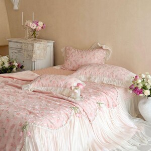 Puede incluir: Un juego de cama rosa con estampado floral de tulipanes, que incluye una colcha y fundas de almohada a juego con ribetes blancos con volantes y peque&ntilde;os lazos de tela decorativos en los bordes, sobre una cama con fald&oacute;n plisado blanco.