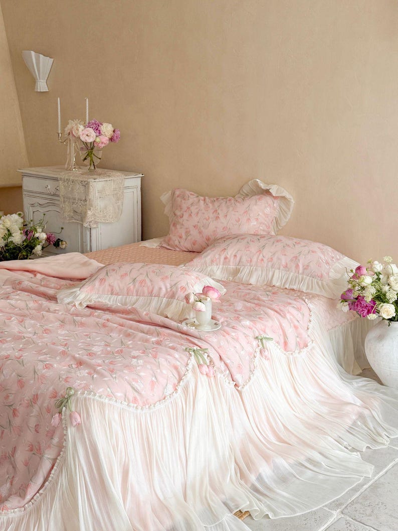 Puede incluir: Un juego de cama rosa con estampado floral de tulipanes, que incluye una colcha y fundas de almohada a juego con ribetes blancos con volantes y peque&ntilde;os lazos de tela decorativos en los bordes, sobre una cama con fald&oacute;n plisado blanco.