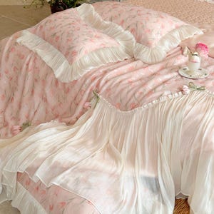 Puede incluir: Un juego de cama de tela rosa suave con un patr&oacute;n de tulipanes repetitivo. El conjunto incluye dos fundas de almohada y una colcha, todas terminadas con amplios volantes blancos plisados en los bordes. La tela tiene una textura ligera.