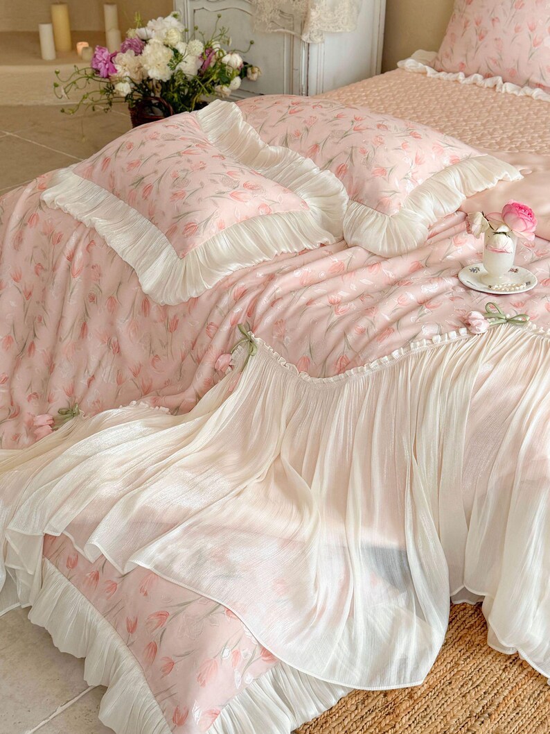 Puede incluir: Un juego de cama de tela rosa suave con un patr&oacute;n de tulipanes repetitivo. El conjunto incluye dos fundas de almohada y una colcha, todas terminadas con amplios volantes blancos plisados en los bordes. La tela tiene una textura ligera.