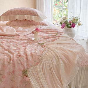 Puede incluir: Un juego de cama rosa claro con un patr&oacute;n repetitivo de tulipanes, que incluye dos almohadas con volantes y una colcha con un borde ancho de tela blanca plisada y transparente. La colcha tiene peque&ntilde;os tulipanes de tela en el borde.
