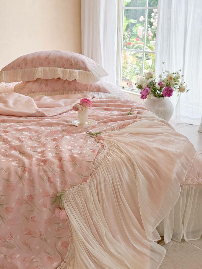 Puede incluir: Un juego de cama rosa claro con un patr&oacute;n repetitivo de tulipanes, que incluye dos almohadas con volantes y una colcha con un borde ancho de tela blanca plisada y transparente. La colcha tiene peque&ntilde;os tulipanes de tela en el borde.