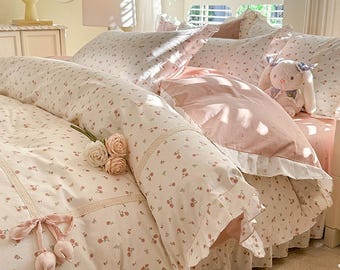May include: A bed dressed with cream and pink floral bedding. The set includes a duvet cover, pillows, and a bed skirt. A stuffed bunny with a blue bow sits on the pillows. A bouquet of roses and a pink ribbon with pom-poms are on the bed.