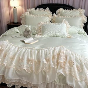 May include: A bed with a cream-colored comforter and matching pillows. The bed skirt features floral embroidery and lace trim. A small purse and an open book rest on the comforter. A lamp is visible in the background.