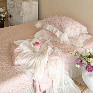Puede incluir: Un juego de cama rosa claro que incluye una colcha acolchada y dos fundas de almohada con bordes de volantes blancos. La tela presenta un suave estampado floral. Un sobrecama de tela plisada blanca a juego est&aacute; colocado sobre la colcha.