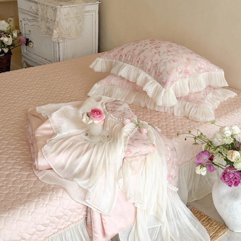 Puede incluir: Un juego de cama rosa claro que incluye una colcha acolchada y dos fundas de almohada con bordes de volantes blancos. La tela presenta un suave estampado floral. Un sobrecama de tela plisada blanca a juego est&aacute; colocado sobre la colcha.