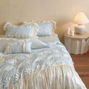May include: A light blue bedding set featuring a white floral applique pattern and ruffled cream-colored trim. The set includes two rectangular pillows with ruffled edges, a smaller rectangular accent pillow, and a duvet cover with a ruffled hem.