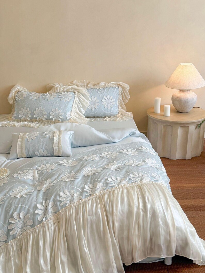 May include: A light blue bedding set featuring a white floral applique pattern and ruffled cream-colored trim. The set includes two rectangular pillows with ruffled edges, a smaller rectangular accent pillow, and a duvet cover with a ruffled hem.