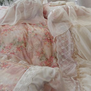 May include: A bed covered with a floral patterned comforter and pillowcases in shades of pink, white, and green. The bedding features delicate lace trim and sheer fabric accents, creating a soft, romantic aesthetic. Two white lace flower appliqués are visible.