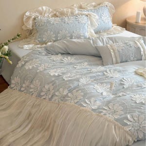 May include: A light blue bedding set featuring a duvet cover and two rectangular pillows with white floral embroidery. The duvet cover and pillows are finished with ruffled cream-colored fabric edges, creating a layered and textured appearance.