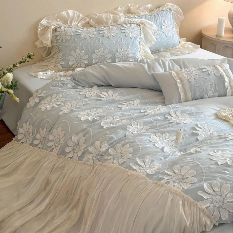 May include: A light blue bedding set featuring a duvet cover and two rectangular pillows with white floral embroidery. The duvet cover and pillows are finished with ruffled cream-colored fabric edges, creating a layered and textured appearance.