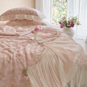 Puede incluir: Un juego de cama rosa claro con un patr&oacute;n repetitivo de tulipanes, que incluye una funda de almohada con volantes y una colcha a juego con un borde de tela blanca plisada. El conjunto est&aacute; decorado con peque&ntilde;os detalles de flores de tela.