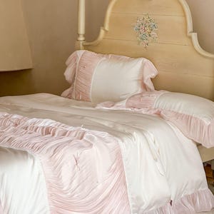 May include: A bedding set featuring a cream-colored duvet cover and pillowcases with soft pink ruffled accents. The fabric has a smooth, satin-like finish, and the ruffles are adorned with small, delicate bows and tiny white beads throughout.