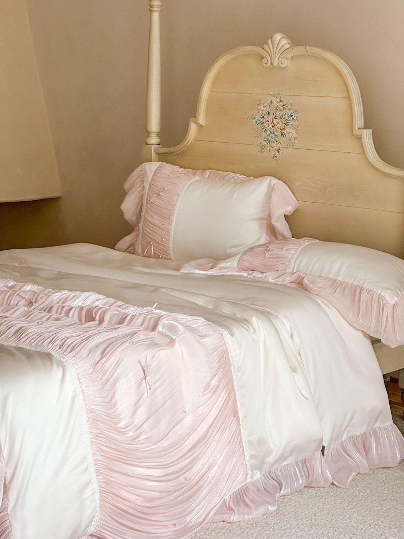 May include: A bedding set featuring a cream-colored duvet cover and pillowcases with soft pink ruffled accents. The fabric has a smooth, satin-like finish, and the ruffles are adorned with small, delicate bows and tiny white beads throughout.