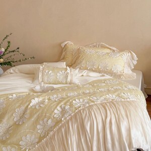 May include: A pale yellow bedding set featuring a duvet cover and pillowcases adorned with white floral applique patterns. The duvet cover includes a wide, pleated white fabric ruffle along the bottom edge, and the pillowcases have ruffled edges.