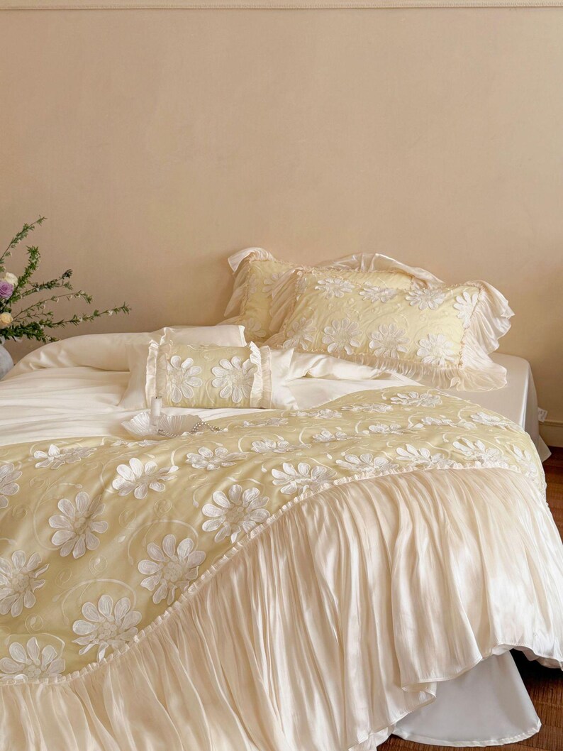 May include: A pale yellow bedding set featuring a duvet cover and pillowcases adorned with white floral applique patterns. The duvet cover includes a wide, pleated white fabric ruffle along the bottom edge, and the pillowcases have ruffled edges.