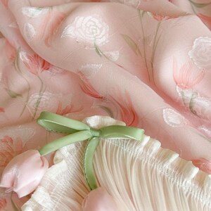 Puede incluir: Una tela rosa claro con un patr&oacute;n de rosas blancas e ilustraciones de tulipanes verde suave. El material est&aacute; fruncido con una cinta verde salvia atada en un lazo, acentuada por dos peque&ntilde;os dijes en forma de tulip&aacute;n rosa p&aacute;lido.