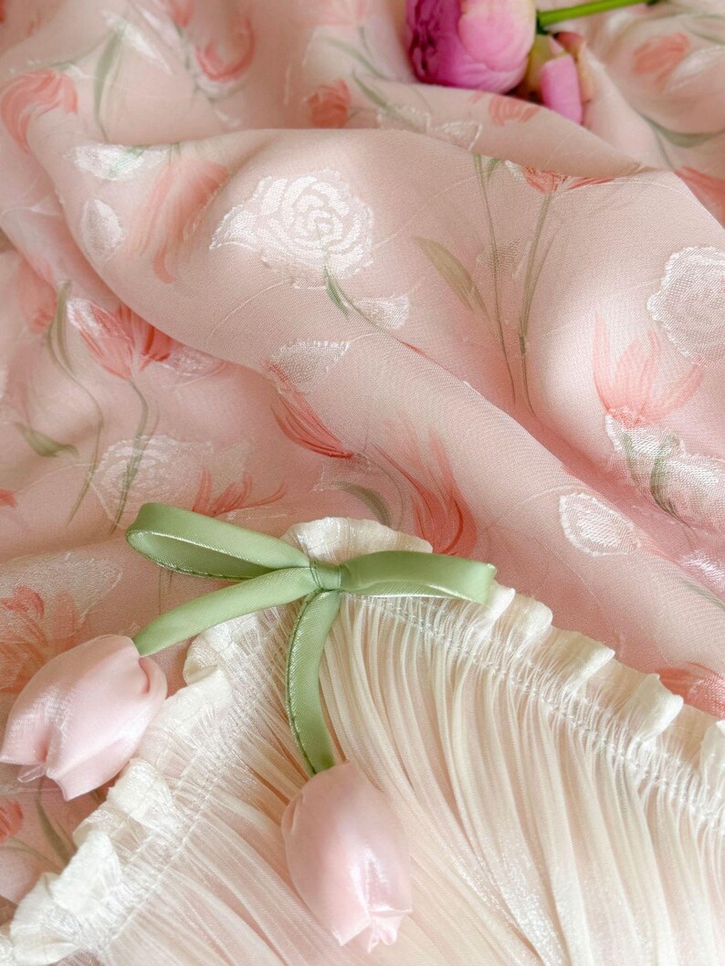 Puede incluir: Una tela rosa claro con un patr&oacute;n de rosas blancas e ilustraciones de tulipanes verde suave. El material est&aacute; fruncido con una cinta verde salvia atada en un lazo, acentuada por dos peque&ntilde;os dijes en forma de tulip&aacute;n rosa p&aacute;lido.