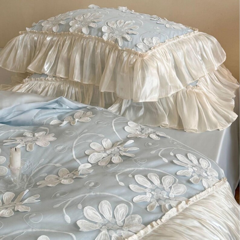 May include: A light blue bedding set featuring a white floral applique pattern and layered ruffled edges. The pillowcase and duvet cover are adorned with embroidered white flowers and vines, finished with wide, gathered cream-colored fabric ruffles.