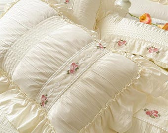 May include: Ivory-colored bedding set featuring ruffled edges and embroidered floral accents. The set includes pillows and a comforter, with a quilted design and delicate pink rose embroidery. The overall aesthetic is soft and romantic.