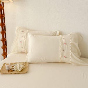 May include: Two off-white pillowcases with embroidered pink floral accents and ruffled edges. The pillows are on a bed with a matching off-white sheet. A wooden tray with a candle and pearl necklace is in the foreground.