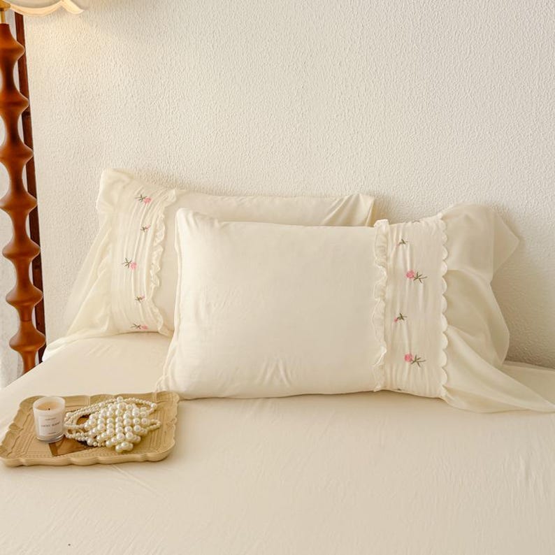May include: Two off-white pillowcases with embroidered pink floral accents and ruffled edges. The pillows are on a bed with a matching off-white sheet. A wooden tray with a candle and pearl necklace is in the foreground.