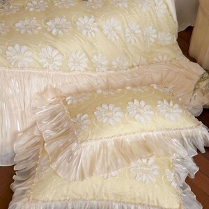 May include: A pale yellow bedding set featuring a duvet cover and two rectangular pillows. The fabric is decorated with white floral appliqu&eacute;s and swirling patterns, finished with wide, translucent, ruffled edges on the pillows and duvet.