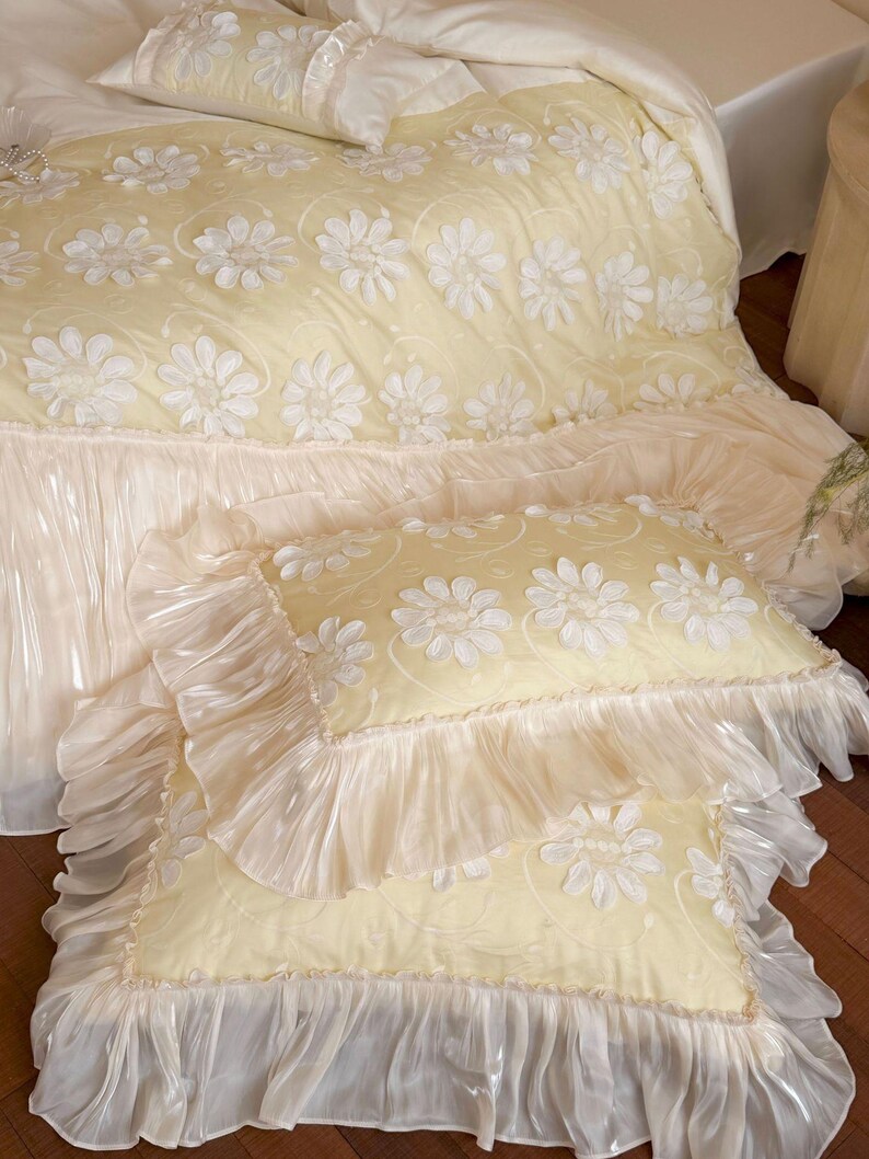 May include: A pale yellow bedding set featuring a duvet cover and two rectangular pillows. The fabric is decorated with white floral appliqu&eacute;s and swirling patterns, finished with wide, translucent, ruffled edges on the pillows and duvet.