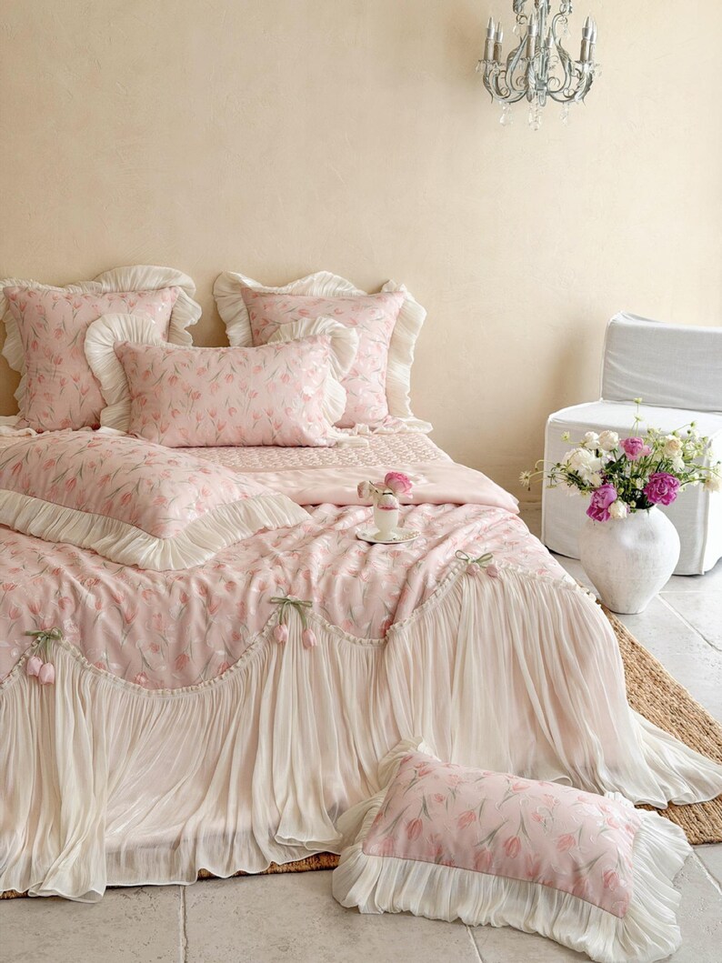 Puede incluir: Un juego de cama de tela rosa claro con un patr&oacute;n repetitivo de tulipanes. El edred&oacute;n y las fundas de almohada est&aacute;n adornados con bordes de volantes color crema y detalles decorativos de tulipanes de tela en el fald&oacute;n.