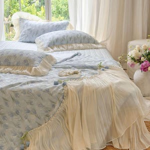 May include: A light blue bedspread and matching pillowcases featuring a repeating tulip pattern. The bedding is accented with cream-colored ruffled fabric trim and small decorative blue tulip embellishments along the edges of the quilt.
