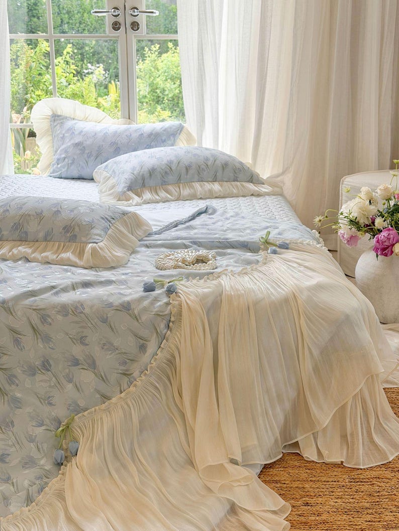 May include: A light blue bedspread and matching pillowcases featuring a repeating tulip pattern. The bedding is accented with cream-colored ruffled fabric trim and small decorative blue tulip embellishments along the edges of the quilt.