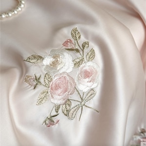 Może przedstawiać: Zbliżenie na jasno różową satynową tkaninę z haftowanym kwiatowym wzorem. Haft przedstawia białe i różowe róże z zielonymi liśćmi i złotymi akcentami. Szereg pereł widoczny jest w lewym górnym rogu.