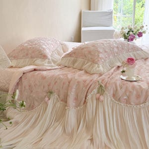 Puede incluir: Un juego de cama rosa claro con un patr&oacute;n de tulipanes repetitivo, dos fundas de almohada a juego con bordes con volantes y una colcha con una falda larga plisada de color crema adornada con peque&ntilde;os tulipanes de tela en el borde.