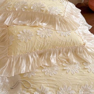 May include: A pale yellow bedding set featuring two rectangular pillows and a matching coverlet. The fabric is adorned with white embroidered floral daisy patterns and finished with wide, gathered ruffles along the edges of the pillows.