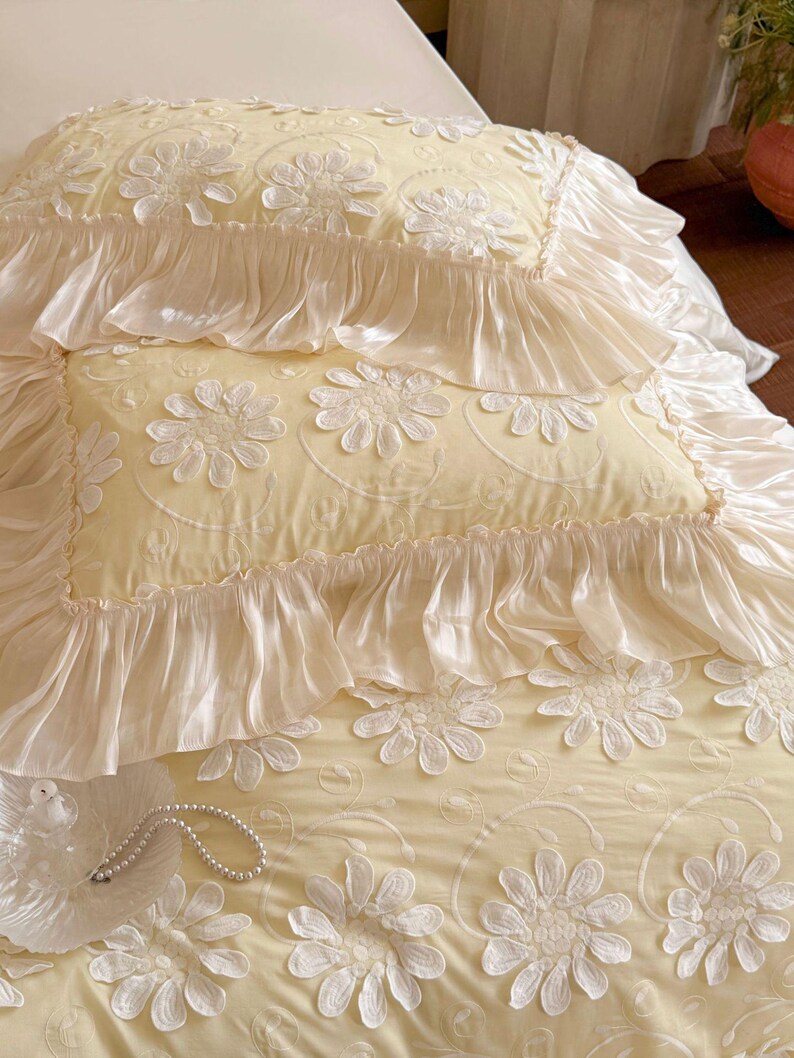 May include: A pale yellow bedding set featuring two rectangular pillows and a matching coverlet. The fabric is adorned with white embroidered floral daisy patterns and finished with wide, gathered ruffles along the edges of the pillows.