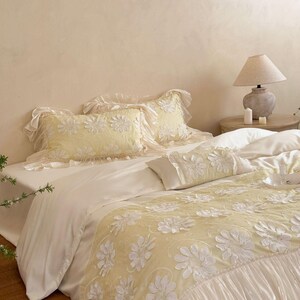 May include: A bedding set featuring a pale yellow duvet cover and matching rectangular pillow shams, all adorned with white floral appliques. The duvet cover includes a wide, ruffled white border at the foot, while the pillows feature delicate ruffled edges.