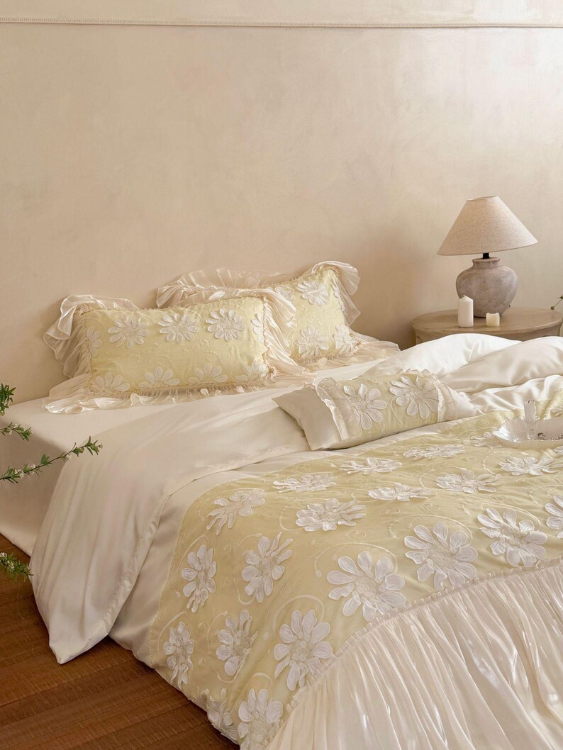 May include: A bedding set featuring a pale yellow duvet cover and matching rectangular pillow shams, all adorned with white floral appliques. The duvet cover includes a wide, ruffled white border at the foot, while the pillows feature delicate ruffled edges.