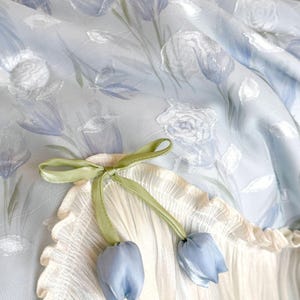 May include: A light blue fabric featuring a white floral pattern with a cream-colored pleated section. A green ribbon is tied into a bow, with two small blue fabric tulip-shaped ornaments hanging from the ends of the ribbon strings.