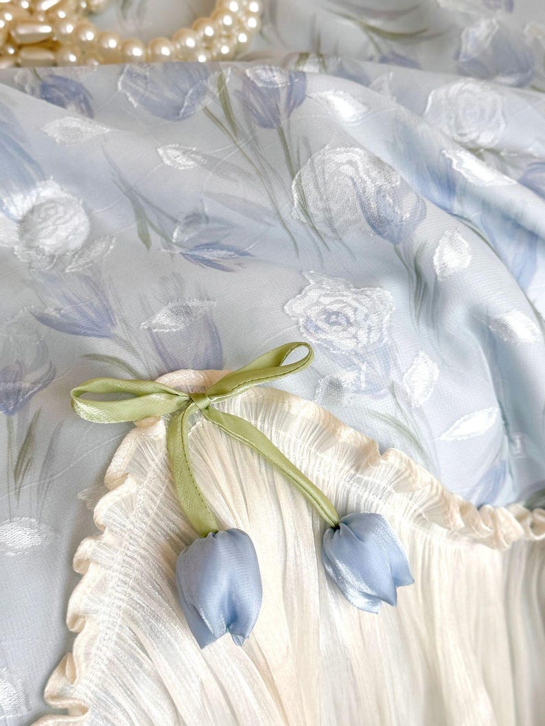 May include: A light blue fabric featuring a white floral pattern with a cream-colored pleated section. A green ribbon is tied into a bow, with two small blue fabric tulip-shaped ornaments hanging from the ends of the ribbon strings.