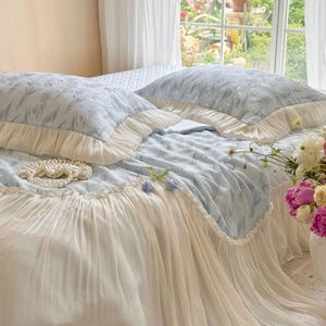 May include: A light blue bedding set featuring a subtle floral pattern, including two rectangular pillows and a bedspread. The pillows and the edge of the bedspread are finished with wide, cream-colored pleated ruffles that drape to the floor.