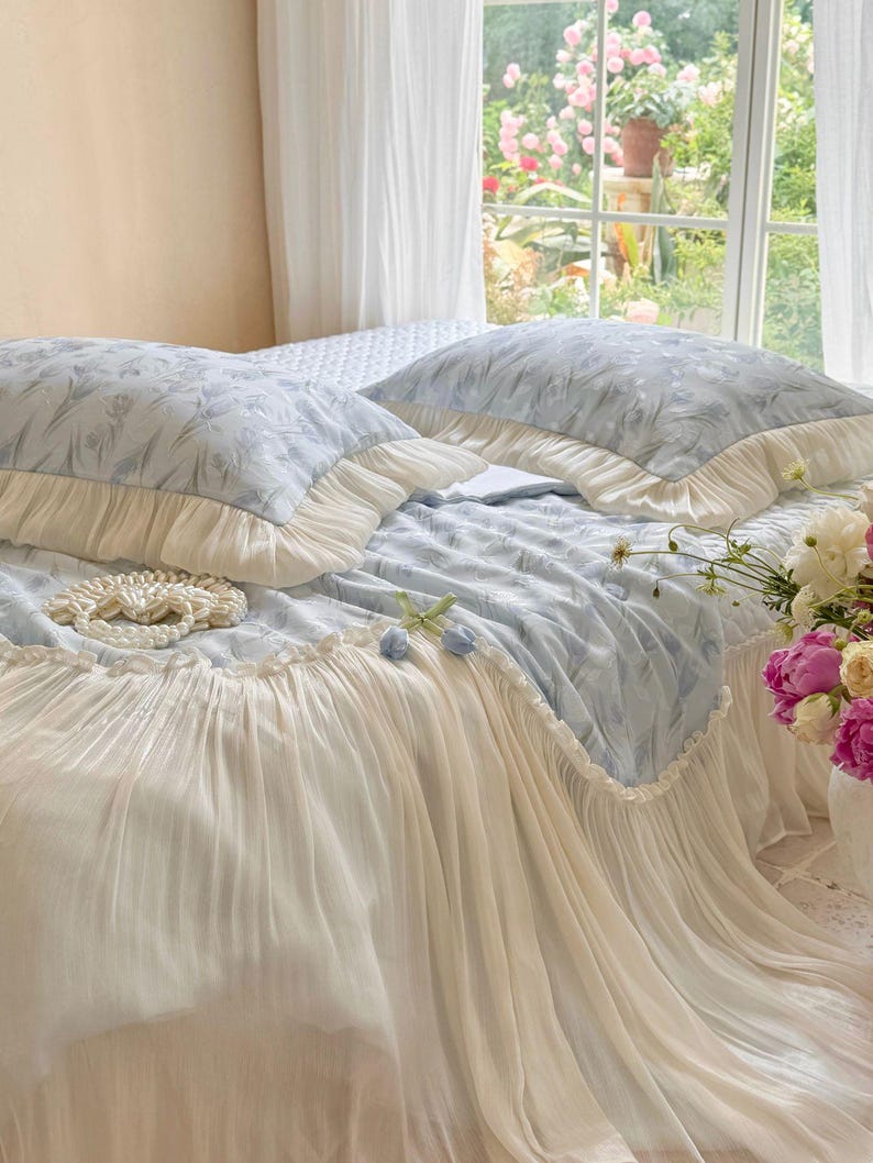 May include: A light blue bedding set featuring a subtle floral pattern, including two rectangular pillows and a bedspread. The pillows and the edge of the bedspread are finished with wide, cream-colored pleated ruffles that drape to the floor.